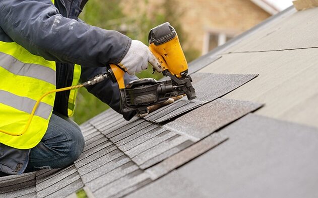 Roofer worker in special protective work wear and gloves, using air or pneumatic nail gun and installing asphalt or bitumen shingle on top of the new roof repairs under construction residential building