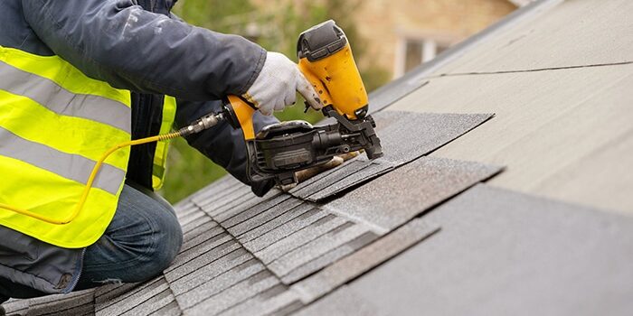 Roofer worker in special protective work wear and gloves, using air or pneumatic nail gun and installing asphalt or bitumen shingle on top of the new roof repairs under construction residential building