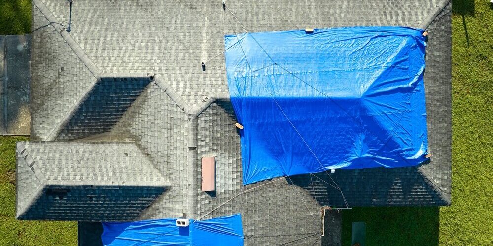 Residential Emergency Roof Repair. Aerial View Of Damaged In storm House Roof Covered. Residential Emergency Roof Repair. Aerial View Of Damaged In storm House Roof Covered.