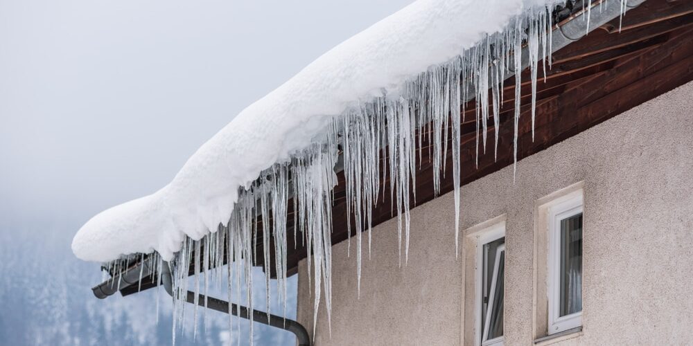 Big,Icicles,And,Snow,Hanging,Over,The,Rain,Gutter,On Big icicles and snow hanging over the rain gutter on a roof of a traditional wooden house in the mountains in winter could be dangerous. Prevention and Maintenance for Godfrey Roofing