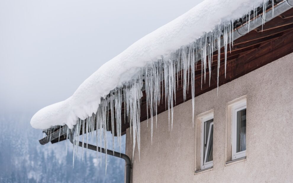 Big icicles and snow hanging over the rain gutter on a roof of a traditional wooden house in the mountains in winter could be dangerous. Prevention and Maintenance for Godfrey Roofing