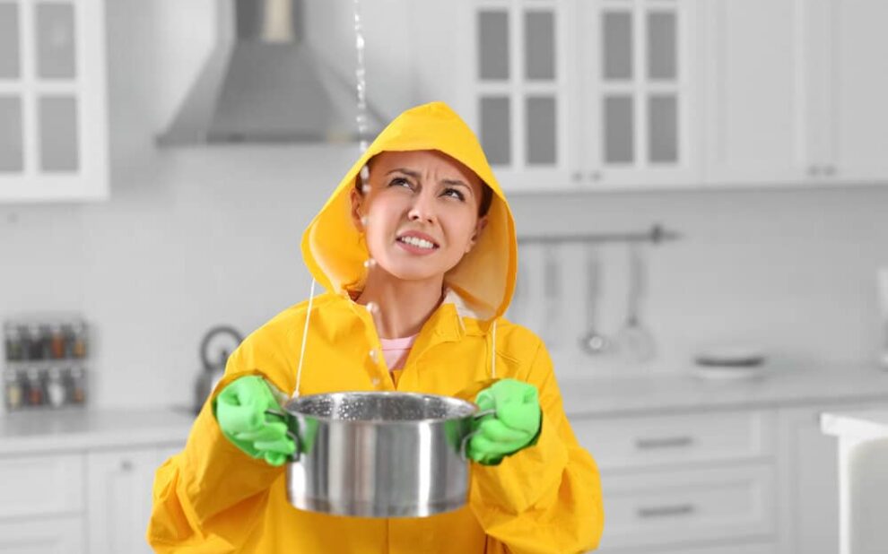Roof Leaks. Young Woman In Raincoat Collecting Leaking Water From Ceiling.