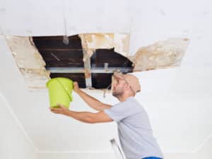 Roof Leaks Structural Damage. Man Collecting Water In Bucket From Ceiling. Ceiling Panels Damaged.