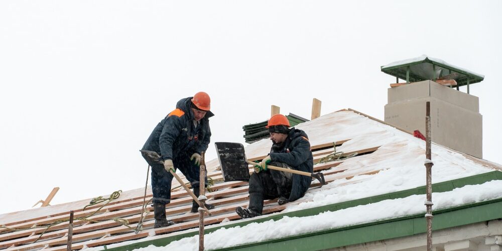 shutterstock_1895989912 (1) Roof Repairs. Roof repairs in winter. Two workers repair the roof on a frosty and snowy day. Men with shovels remove snow from the roof.