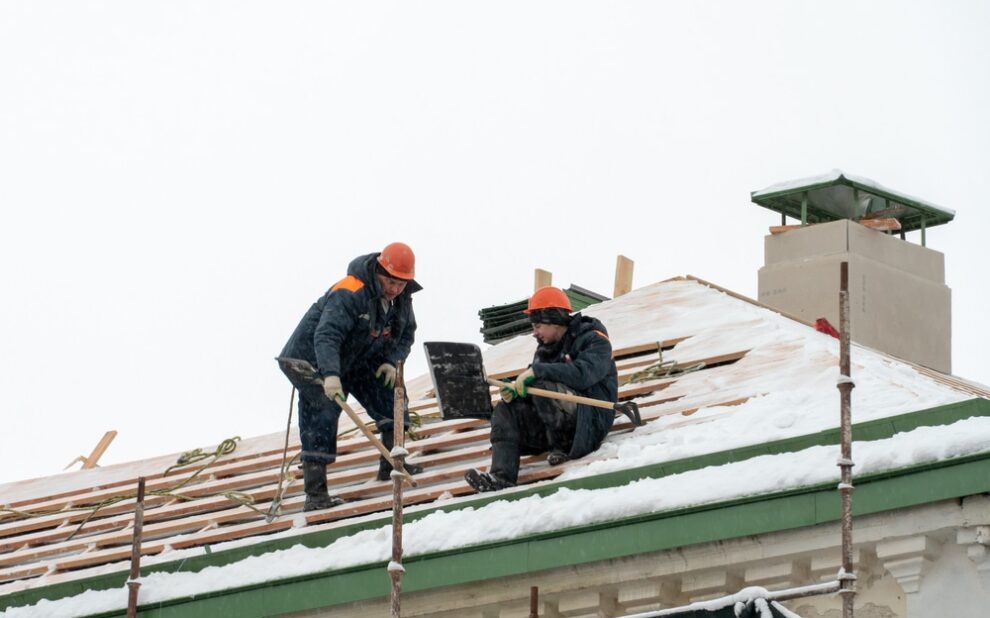 Roof Repairs. Roof repairs in winter. Two workers repair the roof on a frosty and snowy day. Men with shovels remove snow from the roof.