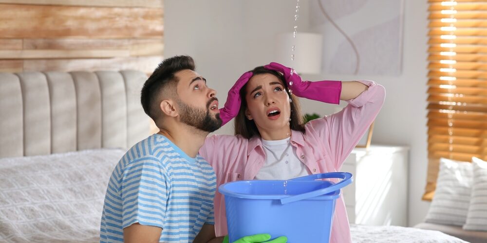 shutterstock_1991456336 (1) Leak Investigation Services. Young couple collecting leaking water from ceiling in bedroom. Time to call roof repair service.