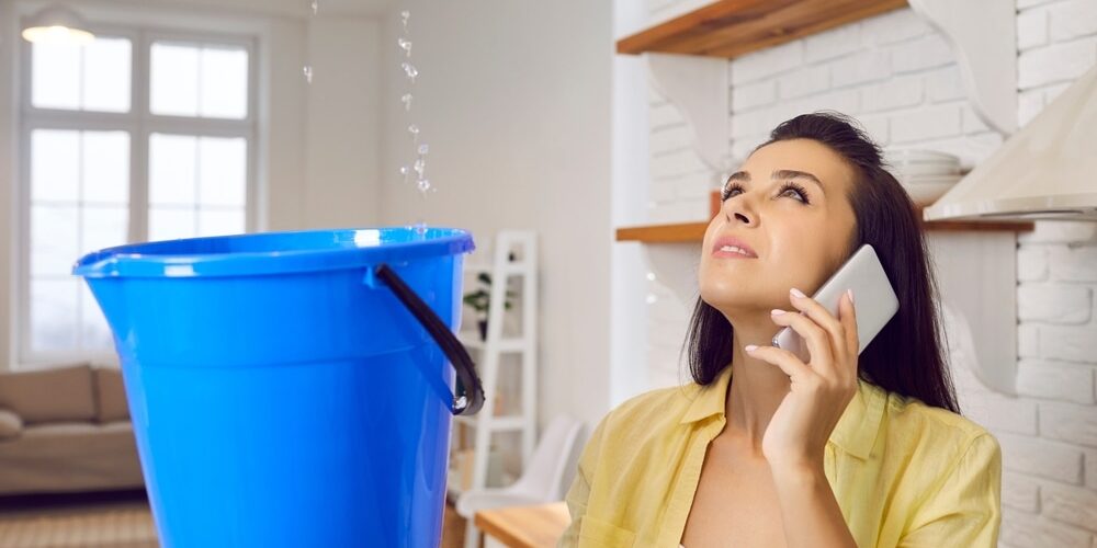 shutterstock_2095335499 (1) Leaky Roof. Sad unhappy frustrated young lady speaking to repair service while holding bucket and looking up at water falling down from damaged ceiling in newly renovated kitchen interior. House roof leak concept.