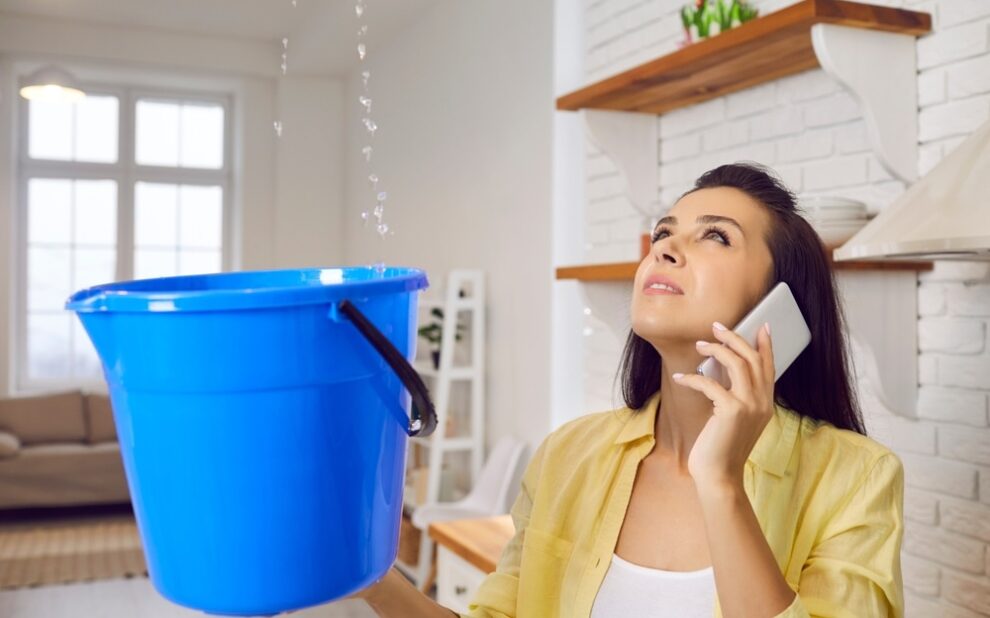 Leaky Roof. Sad unhappy frustrated young lady speaking to repair service while holding bucket and looking up at water falling down from damaged ceiling in newly renovated kitchen interior. House roof leak concept.