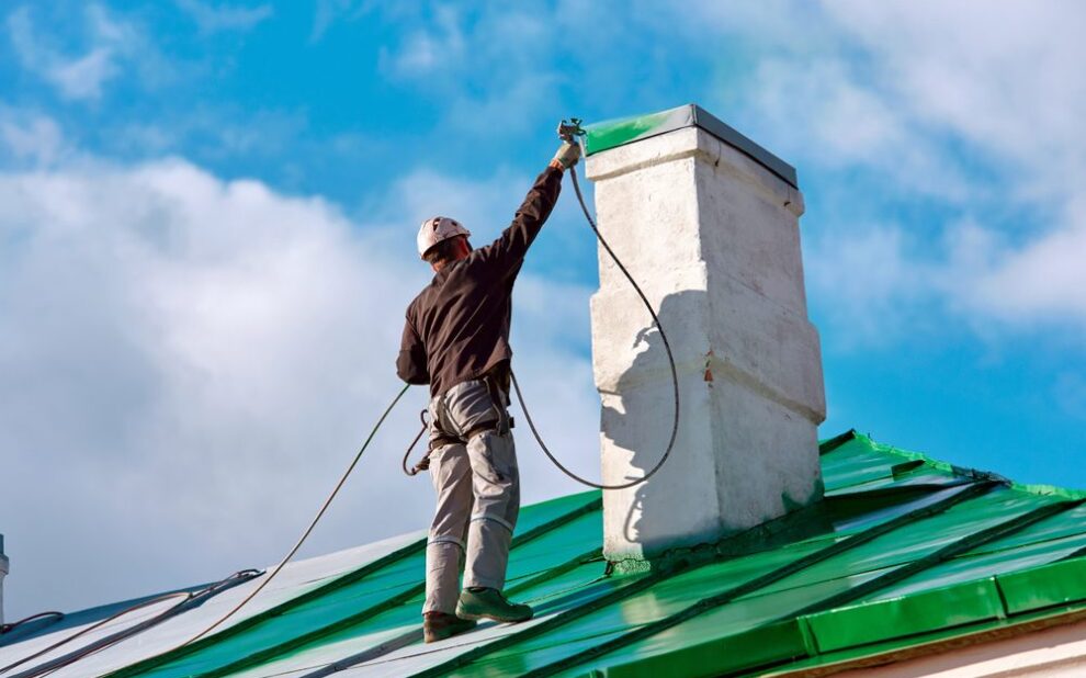 Roof Coatings. Worker of Industrial Alpinist Services painting chimney on the roof with paint spray gun. Professional climber wearing uniform, helmet and using safety harness. Risky job. Extreme occupation.