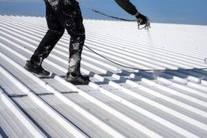 Roof Coatings. Metal roof painting. Technician performing painting of roof in white color. Work at height. Roof repairing service. Rooftop maintenance.