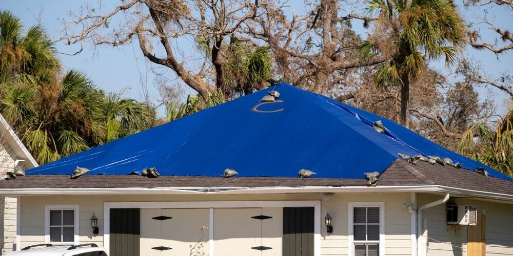 Hurricane,Ian,Damaged,House,Rooftop,Covered,With,Protective,Plastic,Tarp Insurance for Roof Repair. damaged house rooftop covered with protective plastic tarp against rain water leaking until replacement of asphalt shingles. Aftermath of natural disaster.