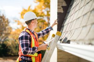 Roof Inspections. A man with a hard hat standing on the steps inspects the house roof.