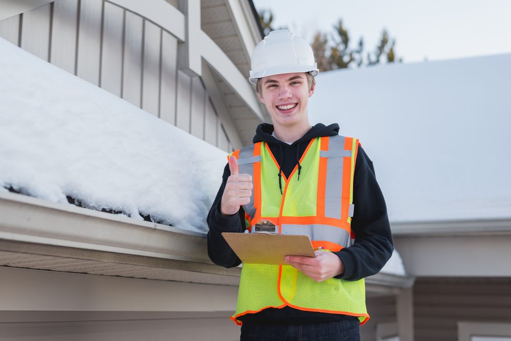 Why Seasonal Roof Inspections Are Important Before Winter Hits. Happy home inspector on a ladder while holding a clipboard and holding thumb up during winter. 