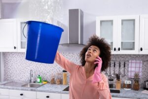 Emergency Roofing Services: What To Do When Your Roof Needs Immediate Repair? Worried Young Woman Calling Plumber On Cellphone And Collecting Leakage Water From Ceiling.