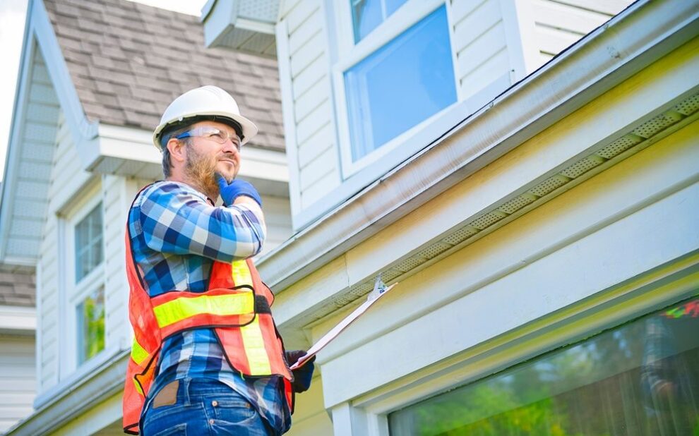 Why Seasonal Roof Inspections Are Critical Before Winter Hits. A man with hard hat standing on steps inspecting house roof.