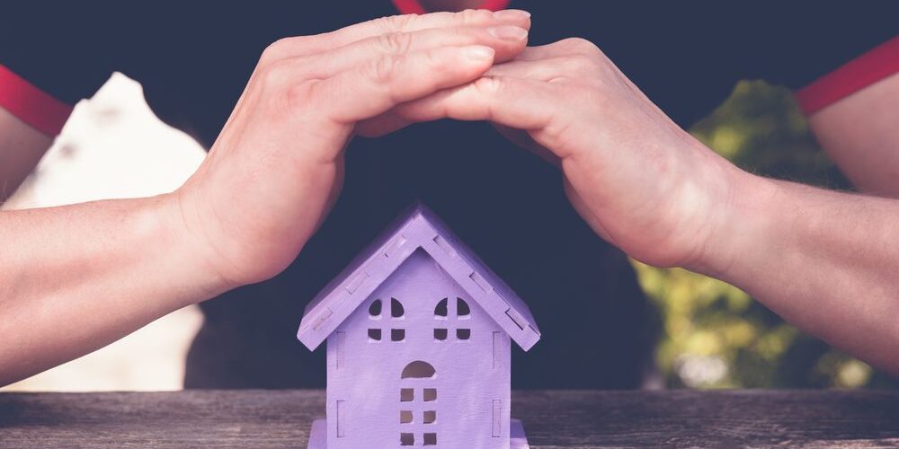 Understanding Your Roof’s Warranty & Winter Damage Coverage. Hands closing a toy house of lavender colour, as a symbol of safety.