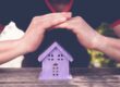 Understanding Your Roof’s Warranty & Winter Damage Coverage. Hands closing a toy house of lavender colour, as a symbol of safety.