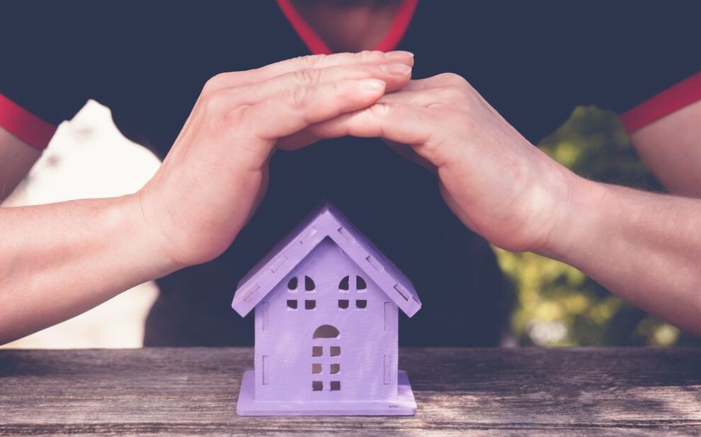 Understanding Your Roof’s Warranty & Winter Damage Coverage. Hands closing a toy house of lavender colour, as a symbol of safety.