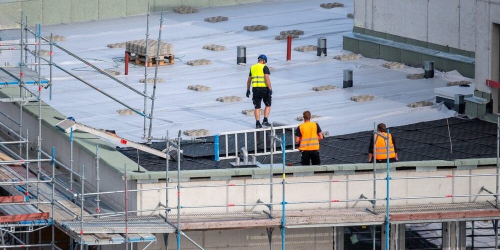 Plan a Commercial Roofing Project Timeline Without Disruption. Birdseye view of roofer waterproofing the flat roof of a commercial building.