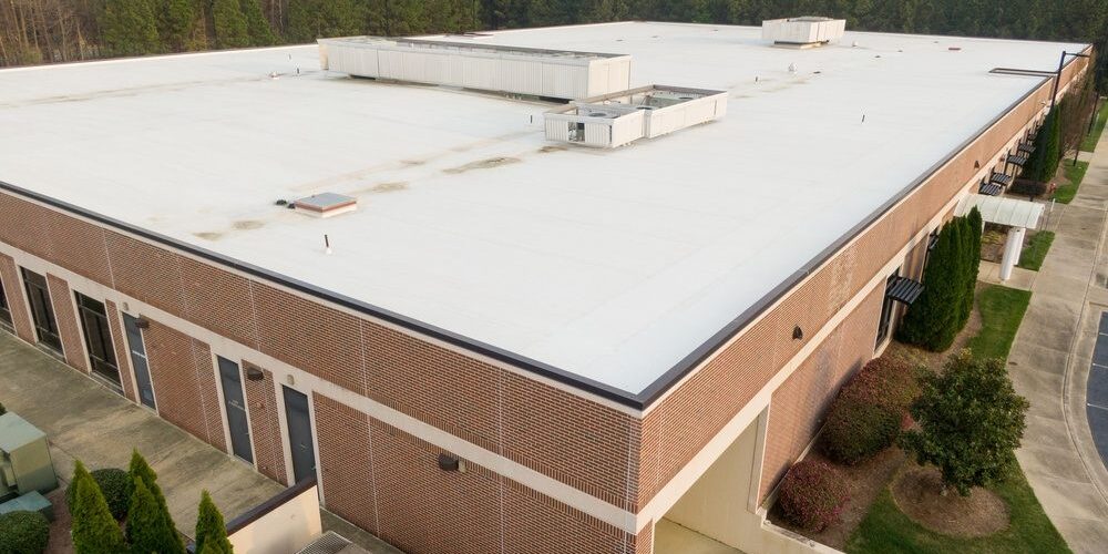 The Step-by-Step Process Commercial Roof Contractors Use for Large-Scale Roof Replacement. Professional aerial perspective of a modern commercial flat roof featuring a white TPO single-ply membrane installation. The image highlights the technical details of the roofing system, including the reflective surface designed for energy efficiency and UV resistance. The shot captures the symmetrical layout of the membrane sheets and the precision of the heat-welded seams, making it ideal for construction industry professionals, roofing contractors, and building inspectors. This high-resolution drone view emphasizes the scale and cleanliness of modern industrial infrastructure and sustainable building practices in 2026.